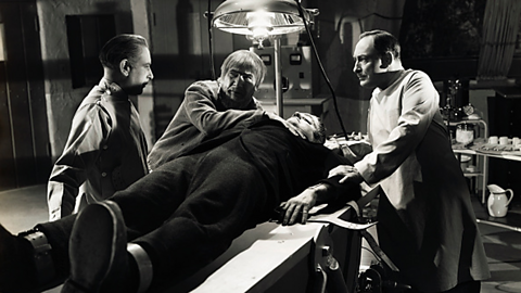 Three men stood around a metal medical bed in a laboratory, with a man with huge feet laying on the bed with his arms folded.