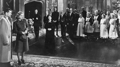 A man and a woman stand in an enormous room with a large fire place surrounded by a large number of unsmiling house staff.