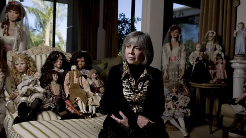 A photograph of a woman dressed in black, sat in a room surrounded by dolls