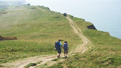 A couple of walkers walking along a costal path that cuts through grass next to the sea below the cliff edge.