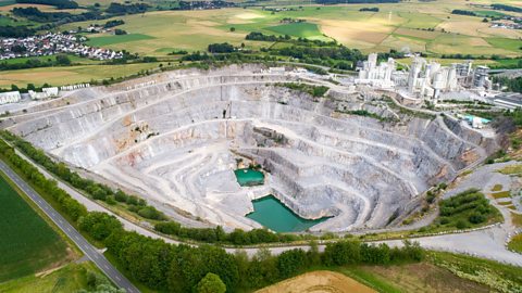 A photo of a large white quarry cut out from the green surroundings. It is a large hole in the rock with buildings to one side.