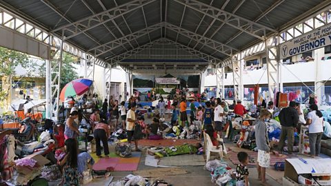 Lots of people under a large sheltered building with no sides surrounded by bedding.