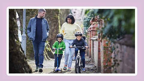 An image of two young boys riding a scooter and a bike with their parents on an pavement in the community with a light purple border around the picture.