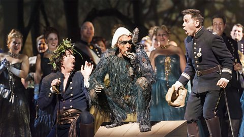 Caliban squats on a wooden box while the people surrounding him look shocked. Caliban is wearing a white piece of cloth on his head, a dark bodysuit designed to make him look hairy and has put something in his mouth.