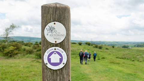 A wooden post at the Antonine Wall on the John Muir Way. A group of walkers can be seen in the background.