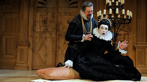 Malvolio kneels behind Olivia with his hands on her shoulders. She has an expression of shock on her face.