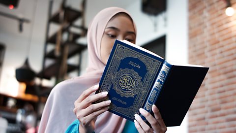 A woman wearing a hijab reads the Quran, which has a blue binding and gold lettering.