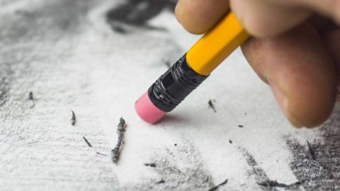 A close-up photograph of a hand rubbing out grey pencil marks with a pencil eraser