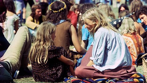 Two young blonde girls wearing baggy clothing sat on a colourful blanket in a crowd of people who are also sat on the grassy floor