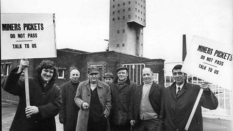 A black and white photo of a group of men standing outside a mine with signs reading 'Miners pickets do not pass talk to us'