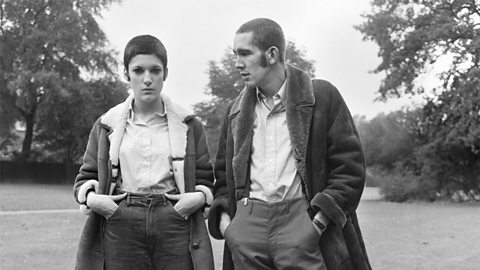 A young man and a young woman standing with their hands in their pockets. They are both wearing long coats and have matching cropped hairstyles.