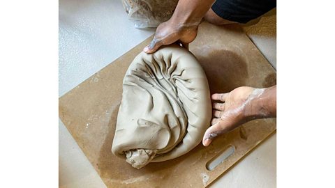 A pair of hands holding a folded mass of wet clay on a wooden board on the floor