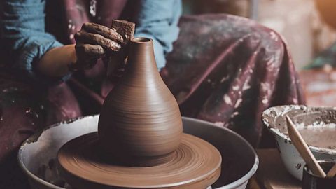 A pair of hands holding a specialist tool and using it to form a vase on a potter's wheel