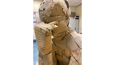 Cardboard sculpture of a figure resting on its elbow with its head on its hand