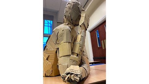 Cardboard sculpture of a human torso and head resting on its elbows on a school desk