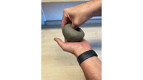 A hand pinching a bowl shape out of a ball of clay with the thumb inside the clay and the fingers outside