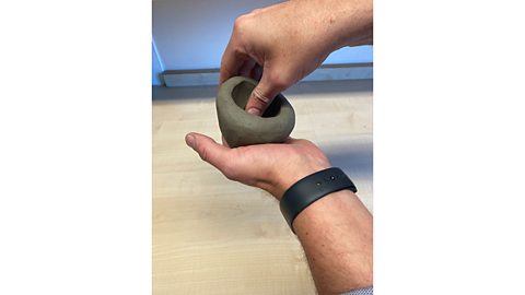 A hand pinching a bowl shape out of a ball of clay with the thumb inside the clay and the fingers outside