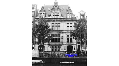 Black and white photograph of a grand house overlooking a river with a blue sports car in colour