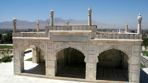 A white marble building with open arches and tall, ornate pillars on the top stands against a background of mountains and blue sky