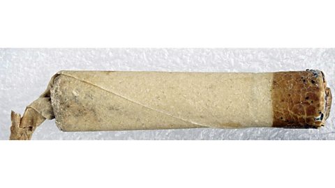 A photo of a long cylindrical object that seems to be wrapped in paper, one end is twisted closed, the other end appears to be dipped in a brown substance. 