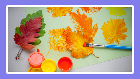 Three leaves and two pots of paint near to a sheet of paper where they have been used to make prints in yellow and orange.