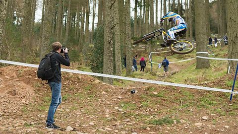 A man photographing a cyclist doing a jump in the woods