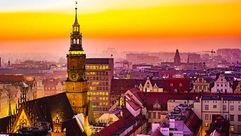 A scene over city rooftops in twilight. A clock tower rising above the rooves