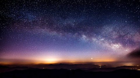 The sky at night. Lots of stars and some hills and lights