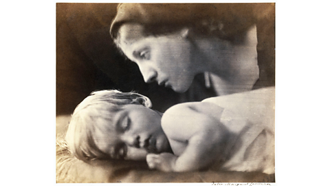 A black and white photograph showing a child lying down asleep, facing the camera, and a mother hovering over him. 