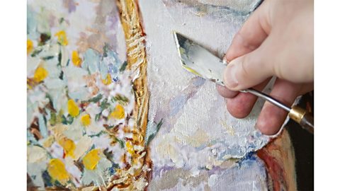 A hand holding a palette knife up to a painting of flowers in a basket.