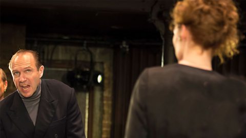 Richard stands with a drink in his hand and an angry expression on his face. Elizabeth stands in front of him.