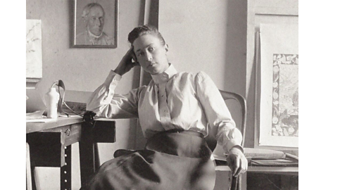 A black and white photo of Hilma Af Klint sitting in a chair, relaxed with her elbow leaning on a table and hand propping up her head.