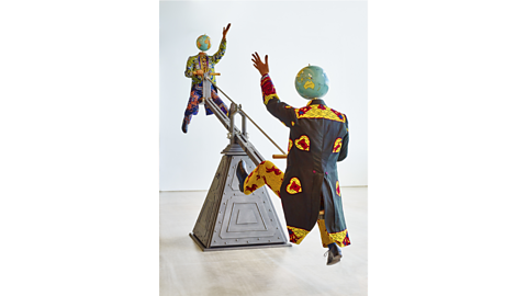 End of Empire installation by Yinka Shonibare, showing two figures dressed in colourful patterned fabric balancing at each end of a rod of metal.
