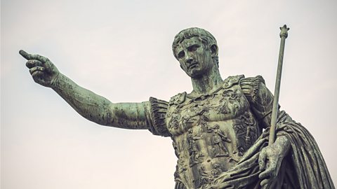 A weathered statue of Augustus, his finger points to the sky. He wears what appears to be an armoured breastplate over his clothes.