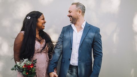 A mature couple celebrating being newly wed. He wears a dark blue suit, she wears a blush pink dress and has a red rose bouquet