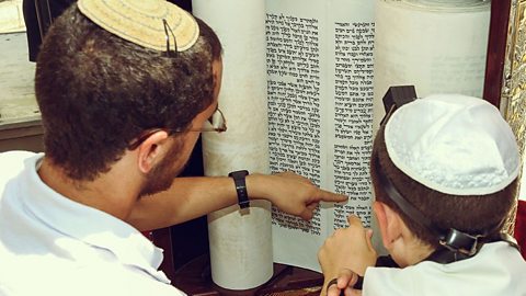 An adult male shows a boy a line from a scroll by pointing at it. Each is wearing a kippah.