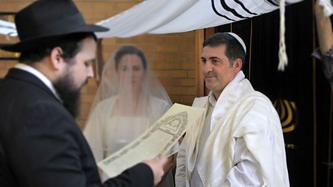 A photo with a rabbi to the left, reading from a scroll, the bride in the middle and groom to the right.