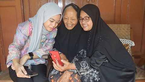 Three generations of women sit together laughing at something on a mobile phone - All are wearing scarves that cover all of their hair.