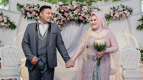 A bride and groom stand hand in hand smiling at each other, the bride wears a scarf covering her hair.