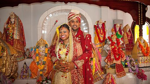 A smiling Hindu couple with his arms around her, both in wedding attire stand  amongst decorated statues of Hindu deities.