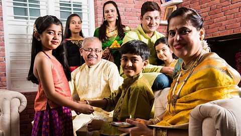 An extended Hindu family, including at least 3 generations (grandparents to grandchildren) sit on a sofa together and smile towards the camera.