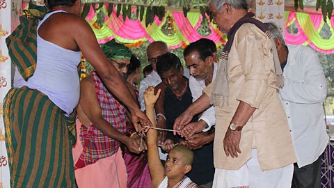 A group of adults stand around a child who is sat on the floor, the child has a shaven head and the adults are helping the child to move their arm through a loop of string that many of the adults are helping to hold.