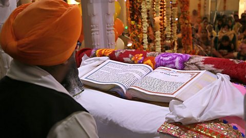 A Sikh man wearing a turban sits in front of a large thick book that is open and laying on a cloth