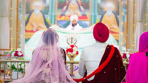 A couple sit on the floor in colourful clothes. A red scarf from the man leads to the woman, who is wearing a pink veil. They sit in front of a large white raised table, at the far end is another Sikh.