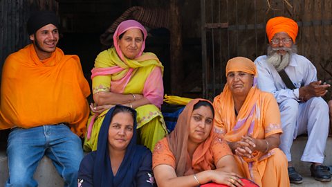 A Sikh family sit together on some steps - The family includes an older grey haired couple, 3 females and a younger male, all smiling.