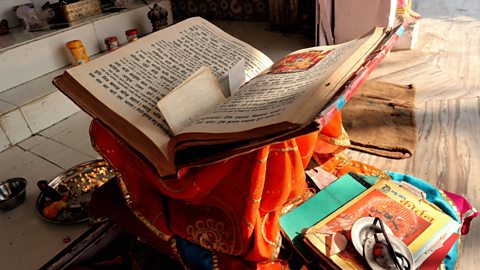 A large book of Hindu scriptures on an altar - There are pieces of note paper in the book and the book is on an orange cloth.