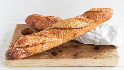 Two baguettes placed on top of each other, one half in a paper bag. Both lying on a wooden chopping board.