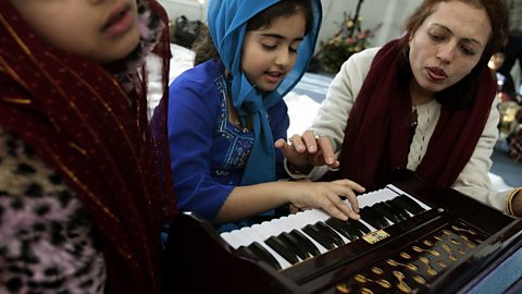 A Sikh woman sits next to a young girl as the girl plays a musical instrument with keys like a piano, called a Harmonium, used in Sikh prayer.