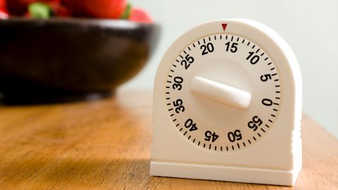 A white cooking timer standing on a wooden counter with the dial pointing at 30 minutes. A bowl of red apples in the background.