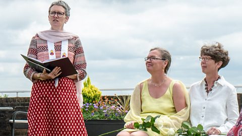 Three people - One person, to the left is standing and reading from a book whilst two women sit down, next to each other to the right, each holding flowers and smiling.